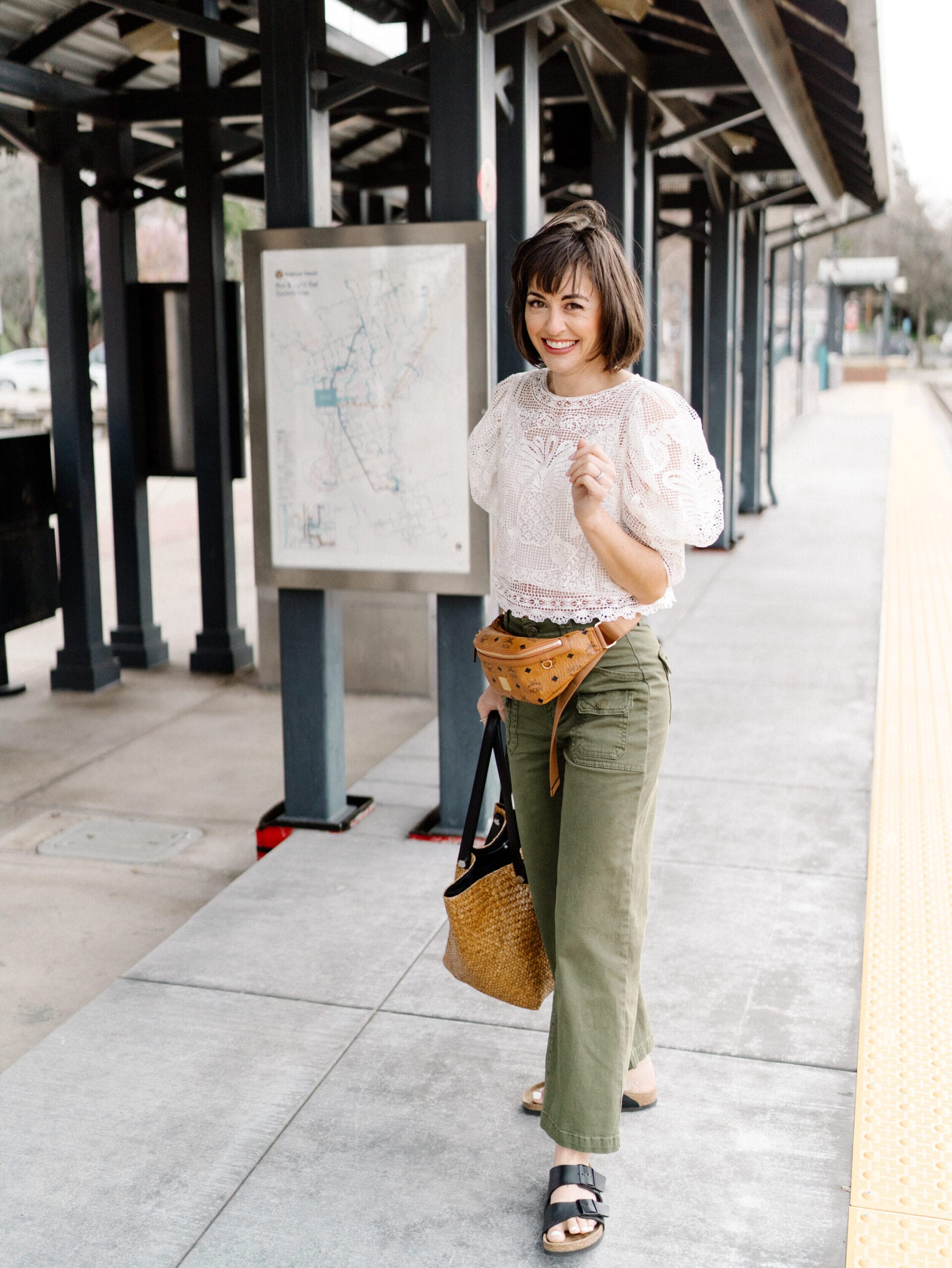 girl at a train station wearing green utility pants from FRAME black birkenstocks and a white lace top mcm brown and black belt bag